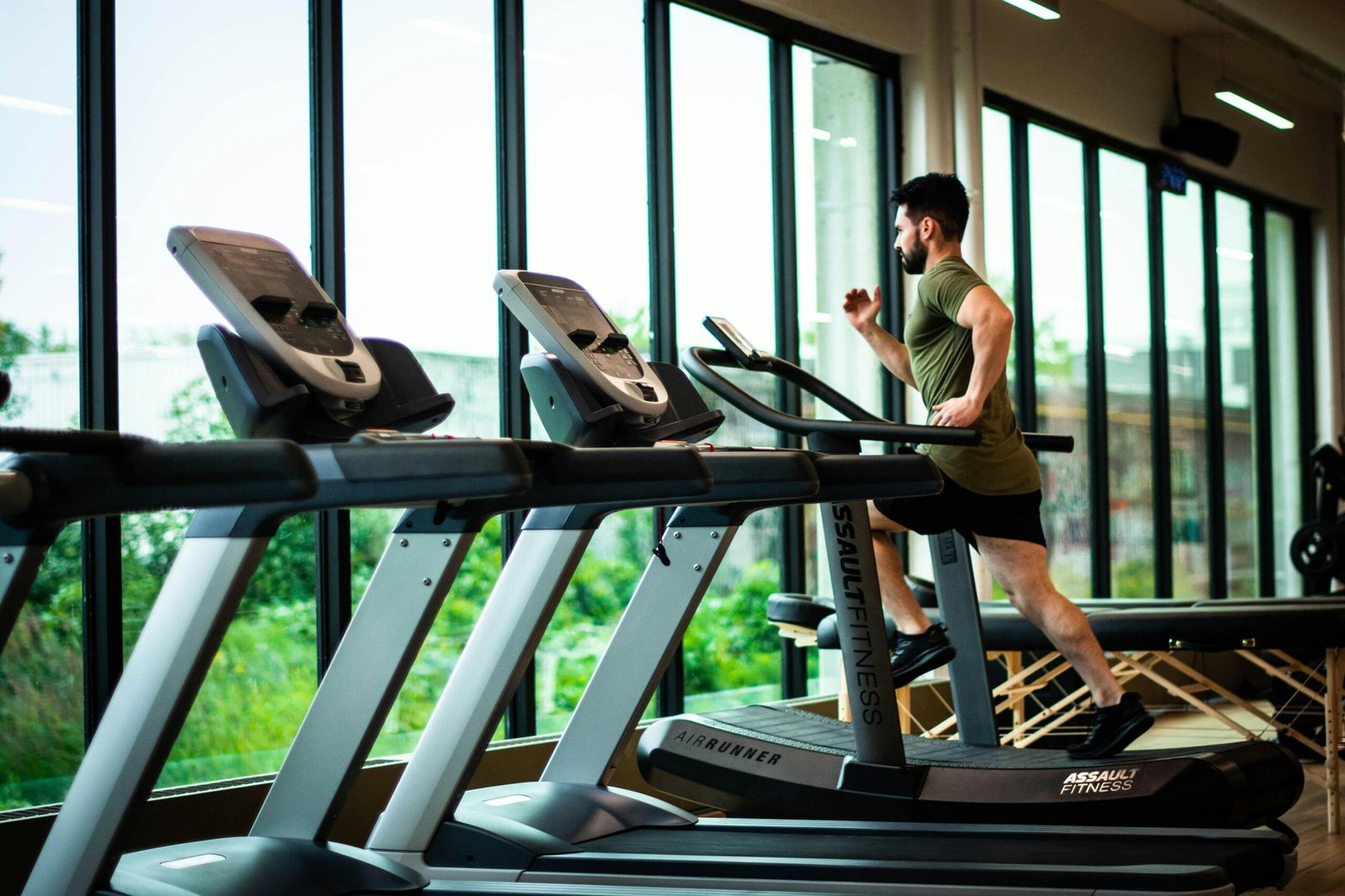 Young man workouts on treadmill in modern gym with large windows and natural light.